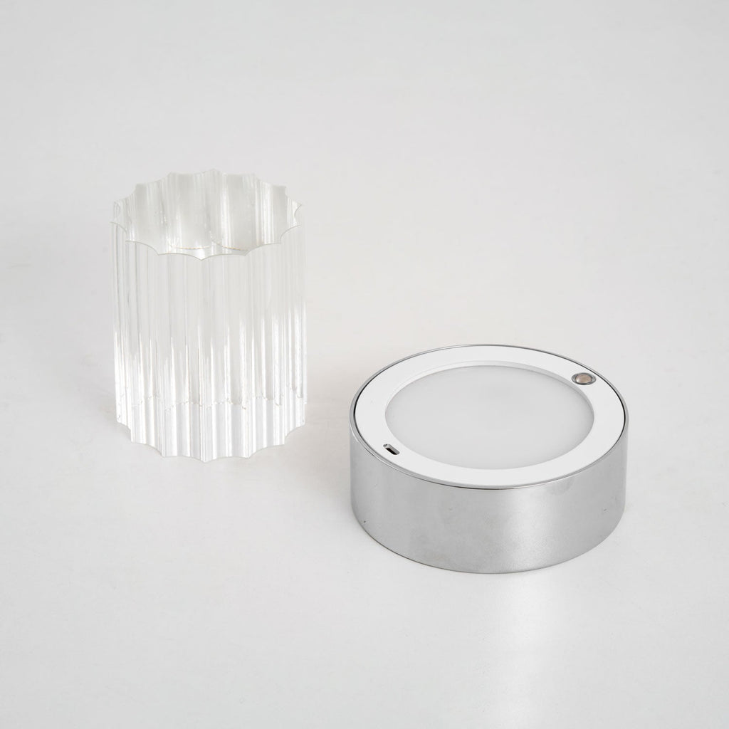 The Clérachic lamp taken apart on a pale surface, with the fluted glass sleeve beside the chrome base and its round light panel.