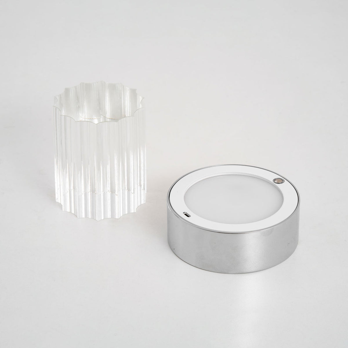 The Clérachic lamp taken apart on a pale surface, with the fluted glass sleeve beside the chrome base and its round light panel.