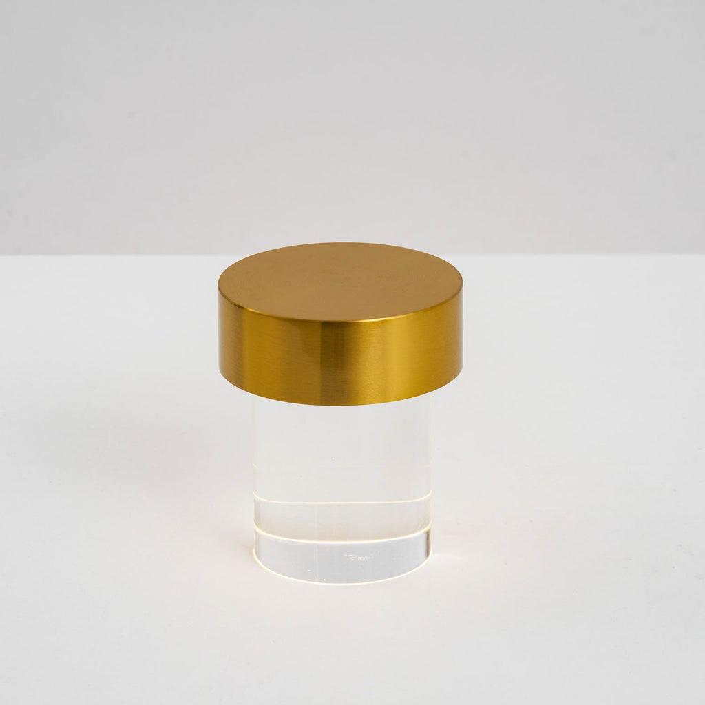 This Clérachic lamp on a pale surface, with a brushed gold cap over smooth clear glass and a ring of light at the foot.