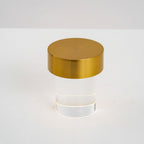 Close view of the brushed gold cap on this Clérachic lamp, with the clear glass cylinder and a thin glowing ring at its base.
