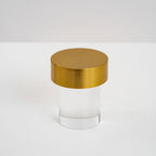 A Clérachic lamp with a brushed gold cap and smooth clear glass on a pale tabletop, unlit.
