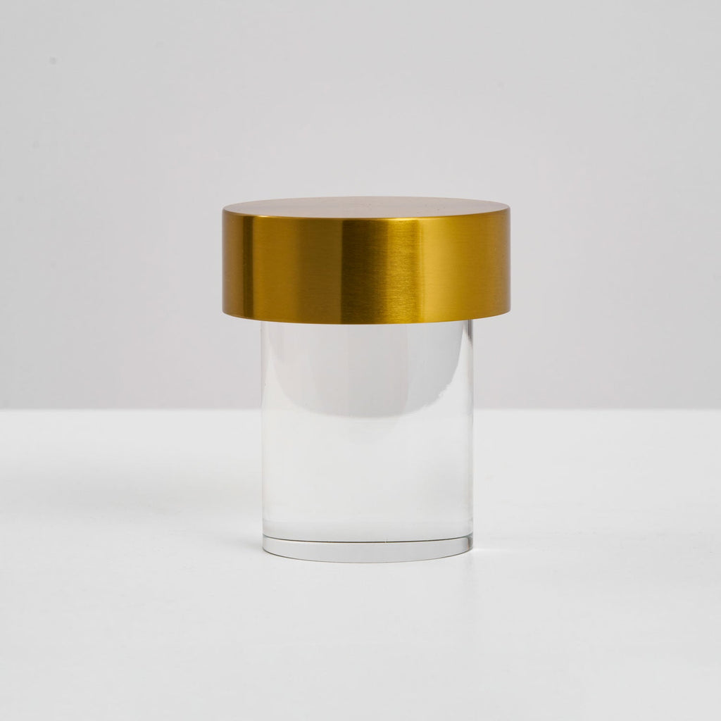 Side view of a Clérachic lamp with a brushed gold cap and a clear glass cylinder, standing on a pale surface.
