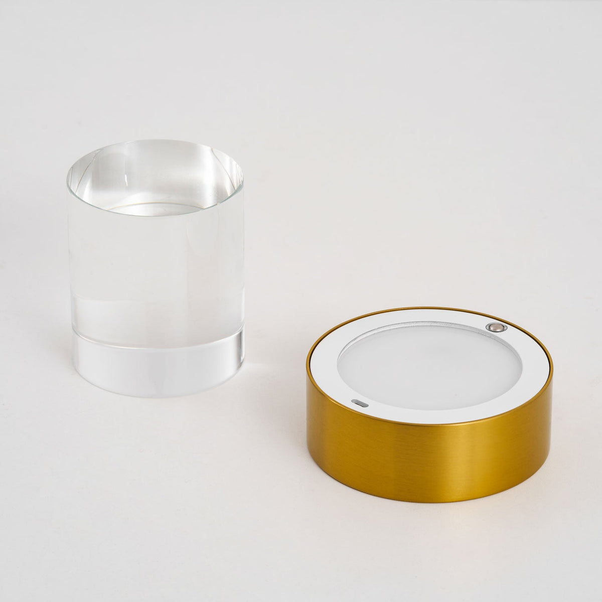 The Clérachic lamp in two parts on a pale surface, with the clear glass cylinder beside the gold base and its round light panel.