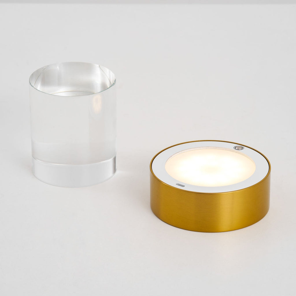 The gold base of this Clérachic lamp switched on, its round panel glowing, beside the separate clear glass cylinder.