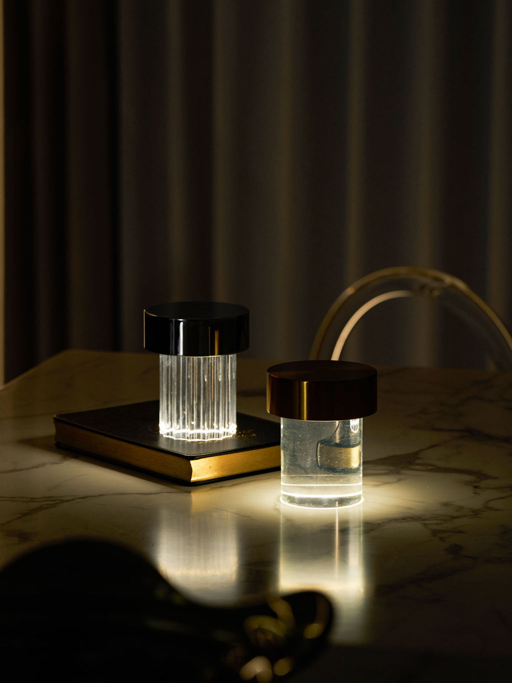Two Clérachic lamps lit on a marble table in a dark room, one fluted with a black cap and one smooth with a dark copper cap.