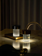 Two Clérachic lamps lit on a marble table in a dark room, one fluted with a black cap and one smooth with a dark copper cap.