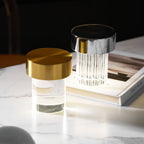 A smooth Clérachic lamp with a gold cap beside a fluted one with a chrome cap, on a marble table with a large book.