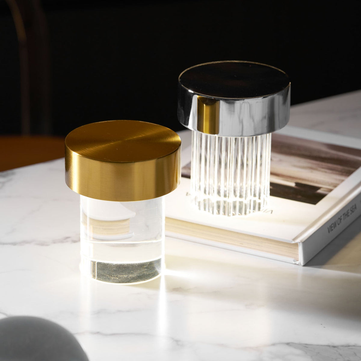 A smooth Clérachic lamp with a gold cap beside a fluted one with a chrome cap, on a marble table with a large book.