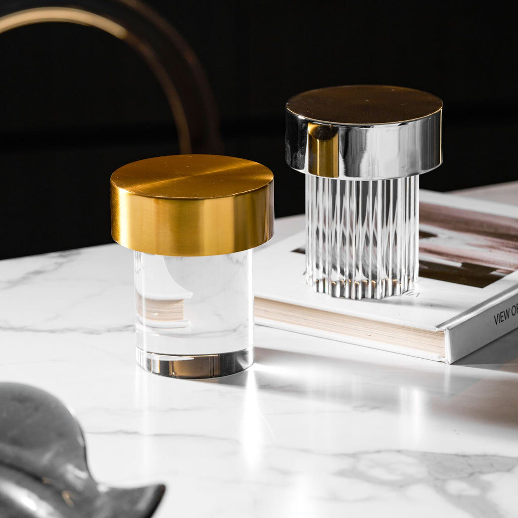 Two Clérachic lamps on a white marble table, one smooth with a gold cap and one fluted with a chrome cap, in a dark room.