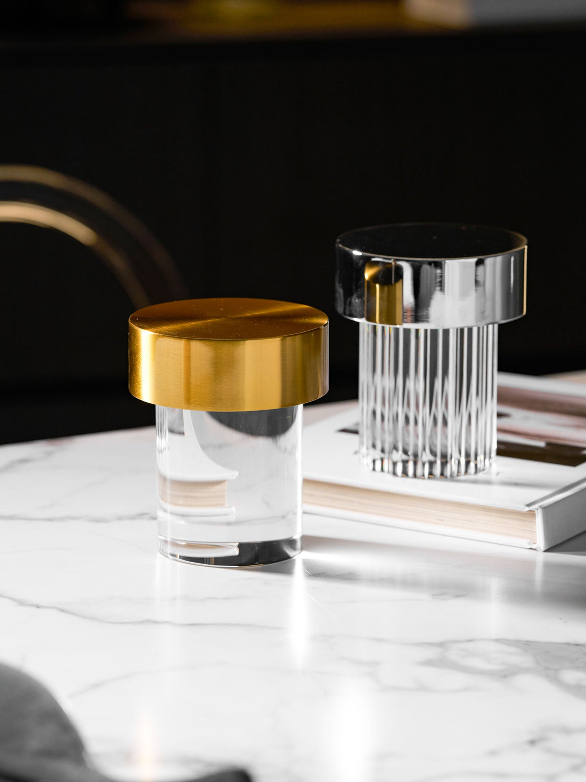 Tall crop of two Clérachic lamps on a white marble table, a smooth gold-capped one in front of a fluted chrome-capped one.