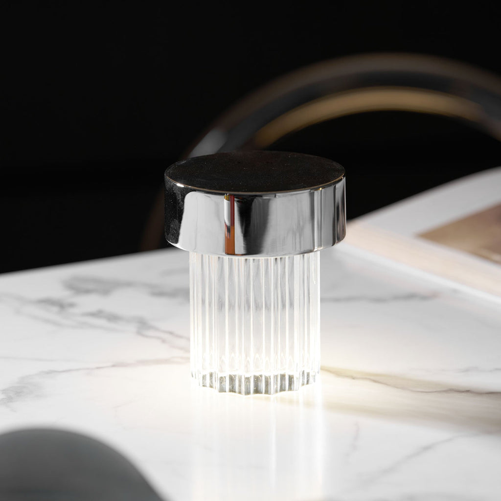 A fluted Clérachic lamp with a chrome cap glowing cool white on a marble table, with a book blurred behind it.