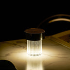 A fluted Clérachic lamp with a dark cap glowing warm on a marble table in a dark room, with a book beside it.