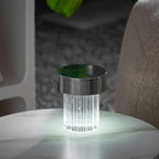 A fluted Clérachic lamp with a chrome cap glowing cool white on a round marble table, with a monstera leaf behind it.