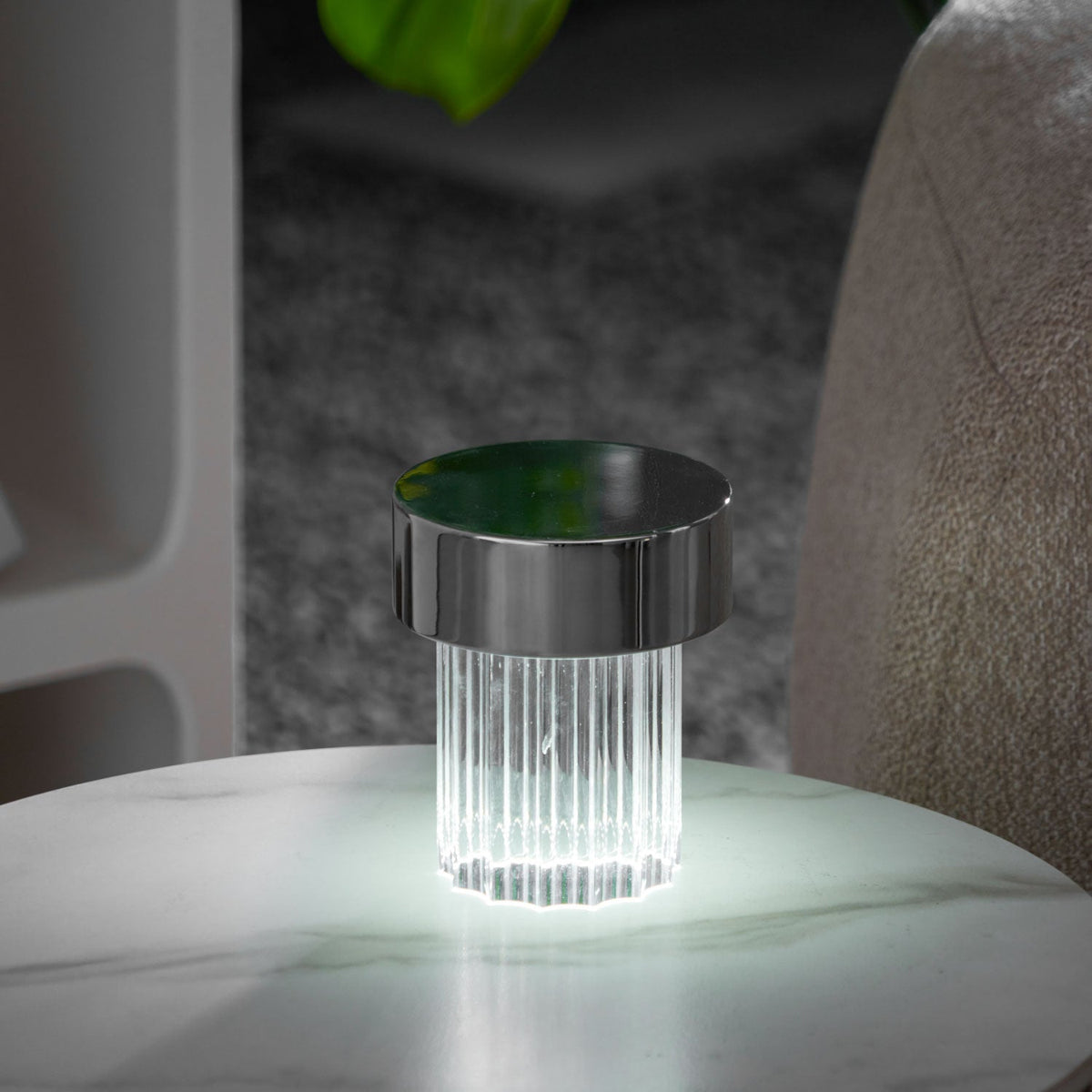 A fluted Clérachic lamp with a chrome cap glowing cool white on a round marble table, with a monstera leaf behind it.