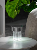 A smaller view of a fluted Clérachic lamp with a chrome cap lit on a round marble table, with monstera leaves above it.