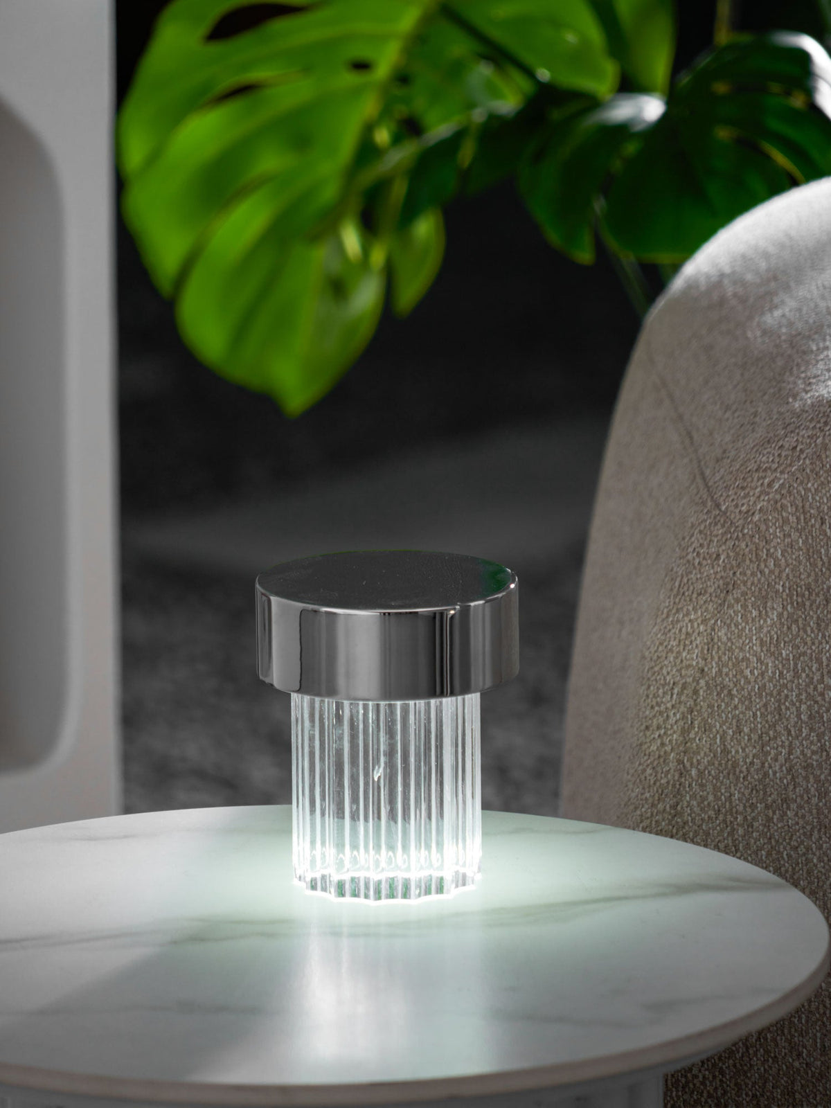 A smaller view of a fluted Clérachic lamp with a chrome cap lit on a round marble table, with monstera leaves above it.