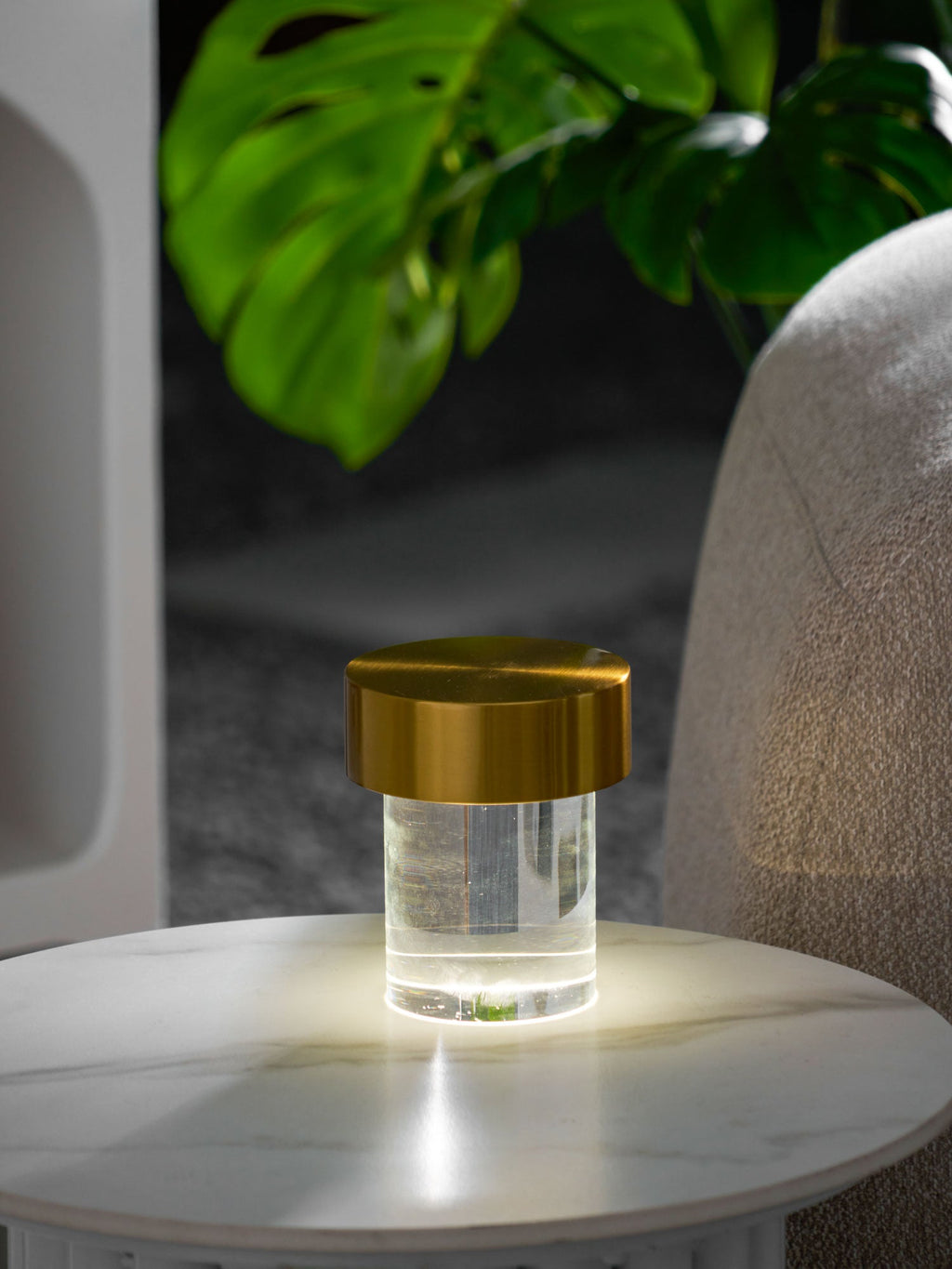 A Clérachic lamp with a gold cap and clear glass glowing warm on a round marble table, with monstera leaves behind it.