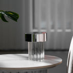 A fluted Clérachic lamp with a chrome cap on a round marble-topped table, with a monstera leaf and grey curtains behind it.