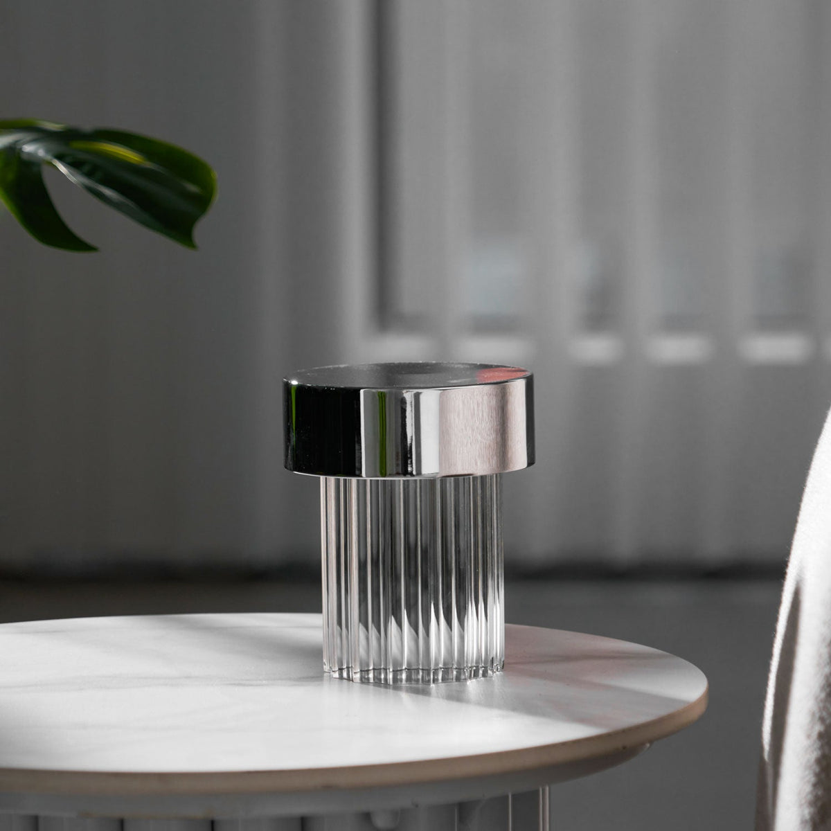 A fluted Clérachic lamp with a chrome cap on a round marble-topped table, with a monstera leaf and grey curtains behind it.