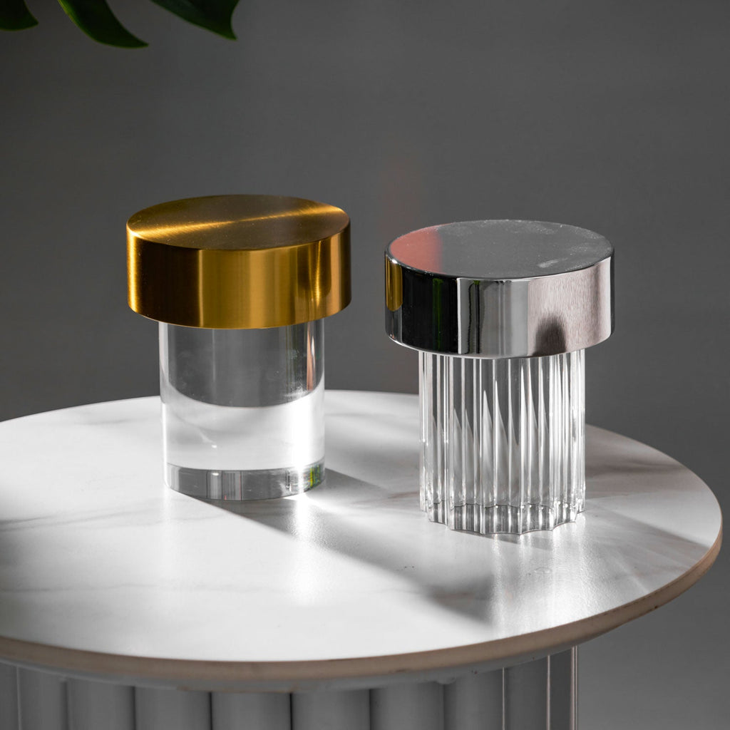 Two Clérachic lamps on a round marble-topped table, a smooth one with a gold cap and a fluted one with a chrome cap.