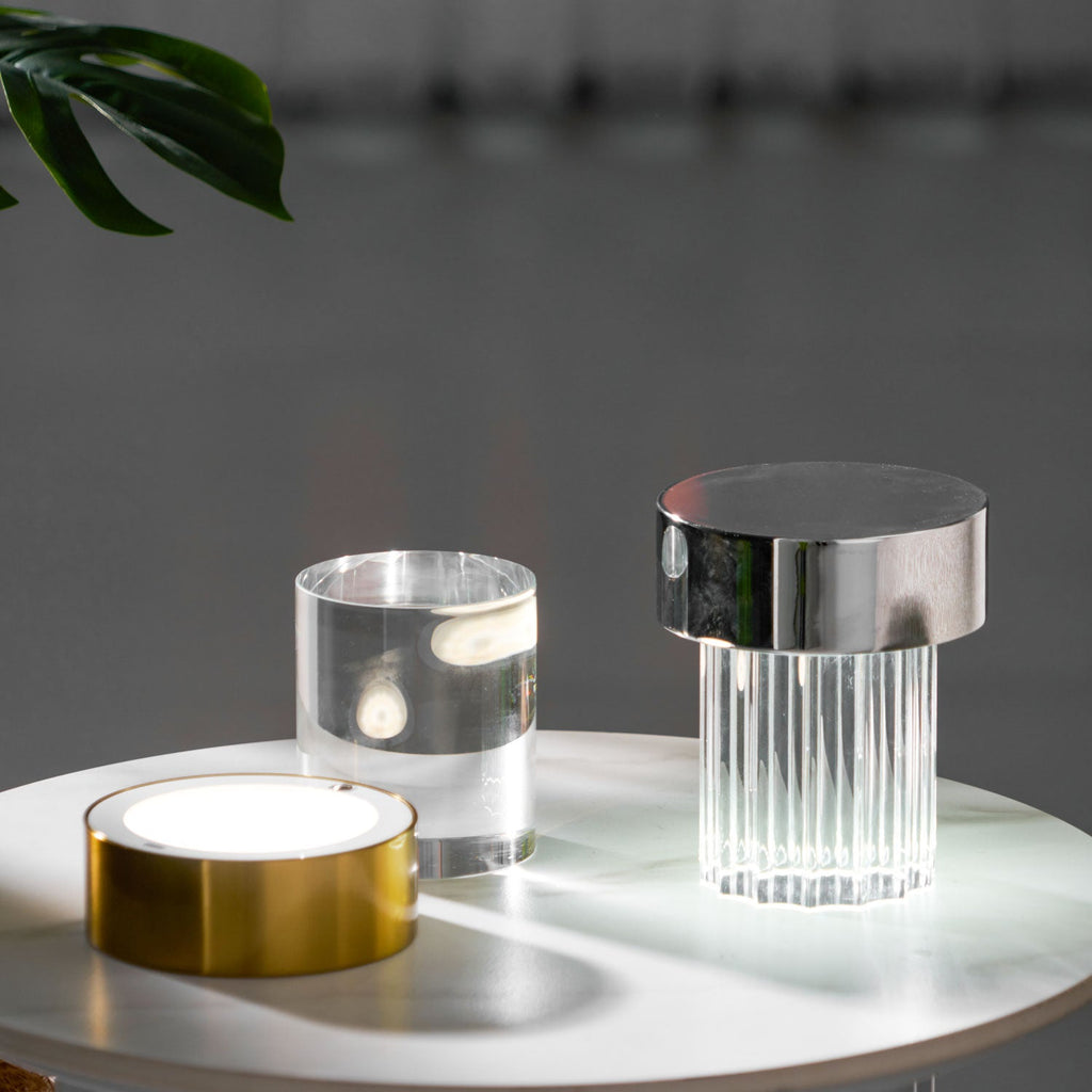 Clérachic lamps on a marble-topped table, with the gold base switched on beside a glass cylinder and a fluted chrome-capped lamp.