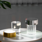 Clérachic lamps on a marble-topped table, with the gold base switched on beside a glass cylinder and a fluted chrome-capped lamp.