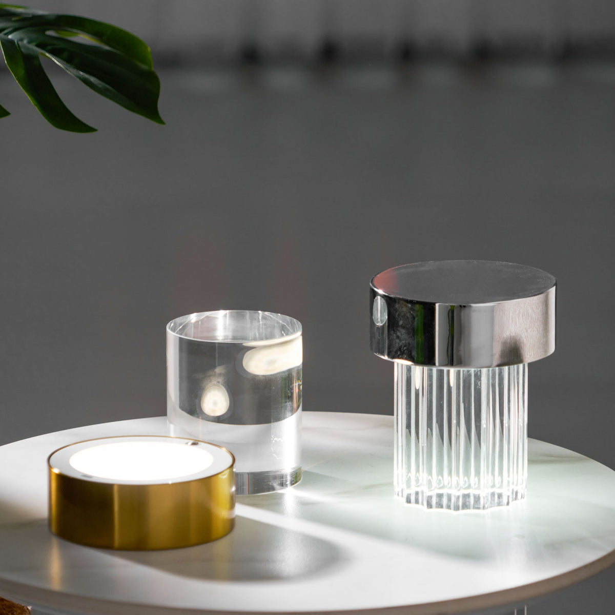 Clérachic lamps on a marble-topped table, with the gold base switched on beside a glass cylinder and a fluted chrome-capped lamp.