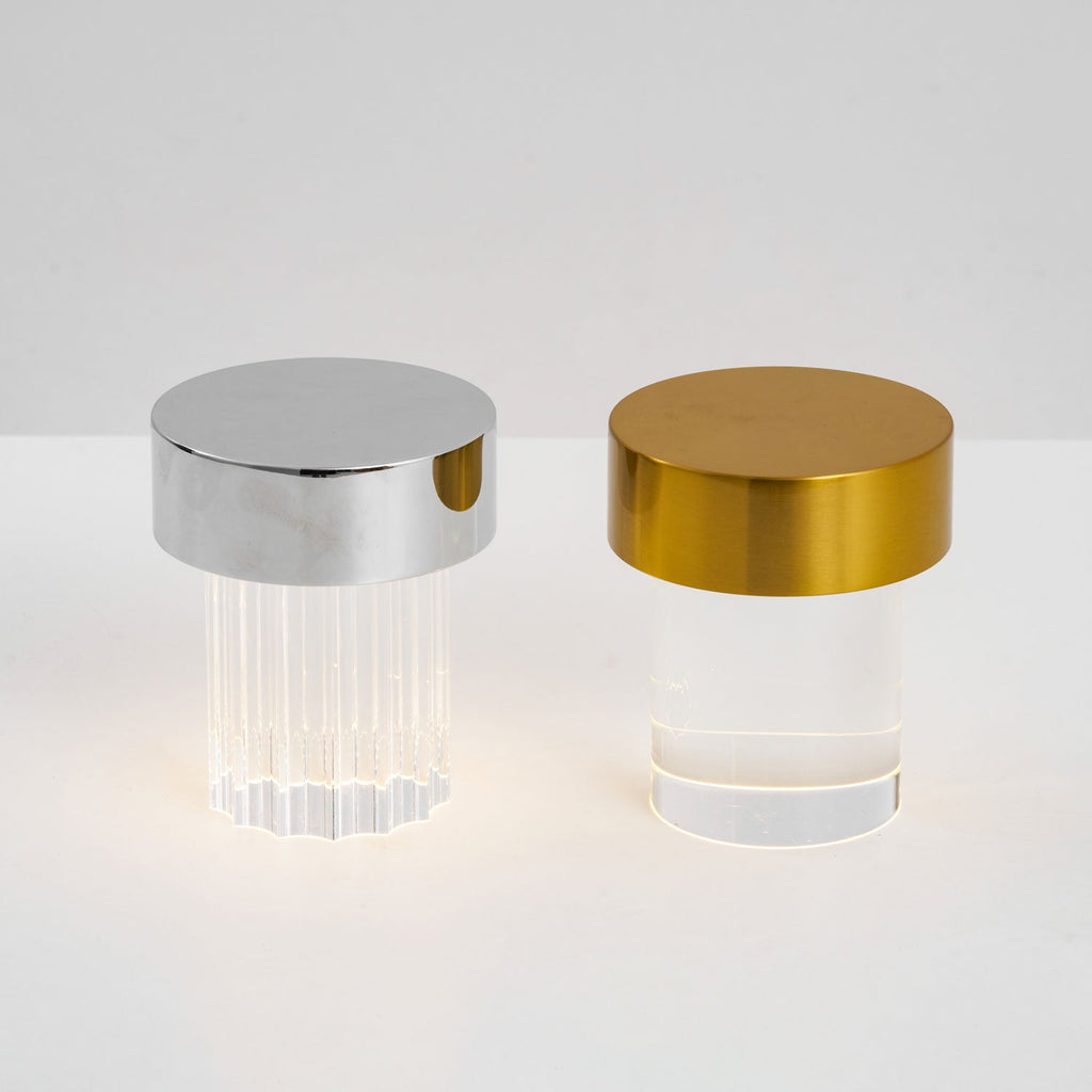 A fluted Clérachic lamp with a chrome cap beside a smooth one with a gold cap, both lit softly on a pale surface.