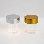 A fluted Clérachic lamp with a chrome cap beside a smooth one with a gold cap, both lit softly on a pale surface.