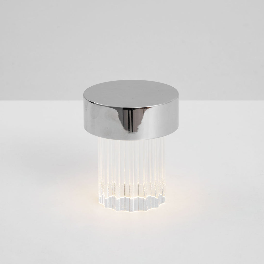 A fluted Clérachic lamp with a chrome cap lit at the base on a pale tabletop, seen from slightly above.