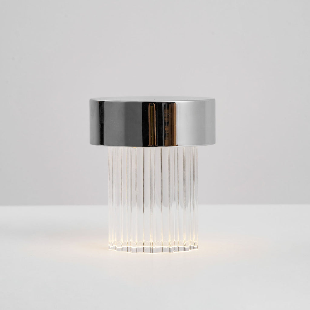 Side view of a fluted Clérachic lamp with a chrome cap, with a faint glow at the base on a pale surface.