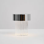 Side view of a fluted Clérachic lamp with a chrome cap, with a faint glow at the base on a pale surface.