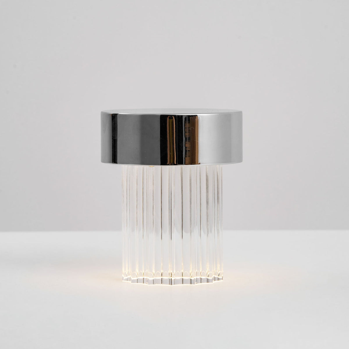 Side view of a fluted Clérachic lamp with a chrome cap, with a faint glow at the base on a pale surface.