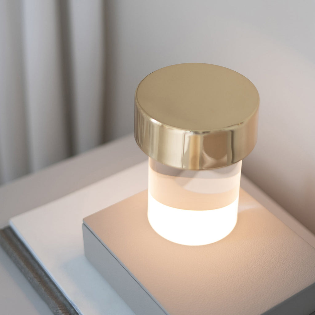 Lit Clérachic table lamp with a gold cap, its lower glass body glowing warm white on a stack of books by a pale wall.