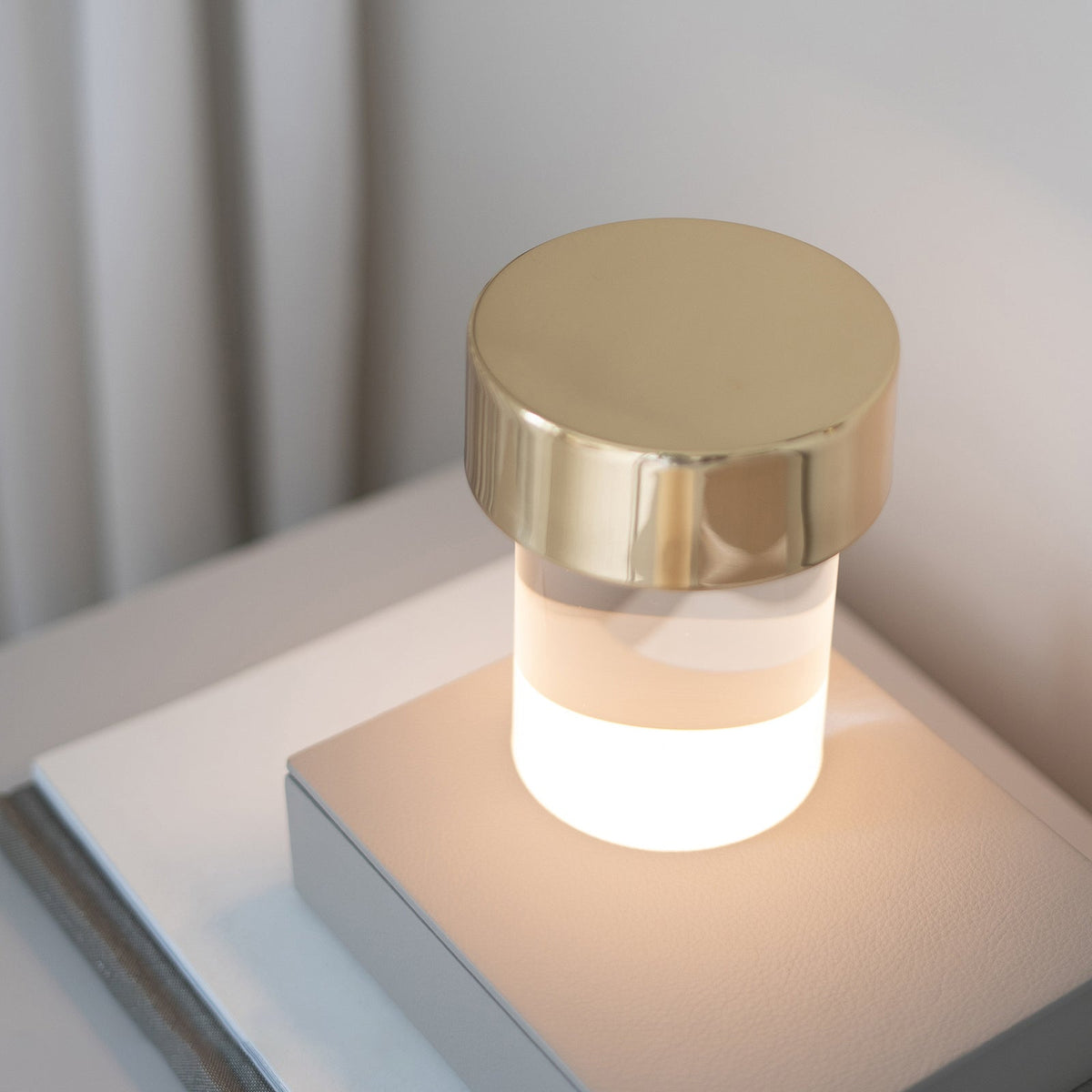 Lit Clérachic table lamp with a gold cap, its lower glass body glowing warm white on a stack of books by a pale wall.