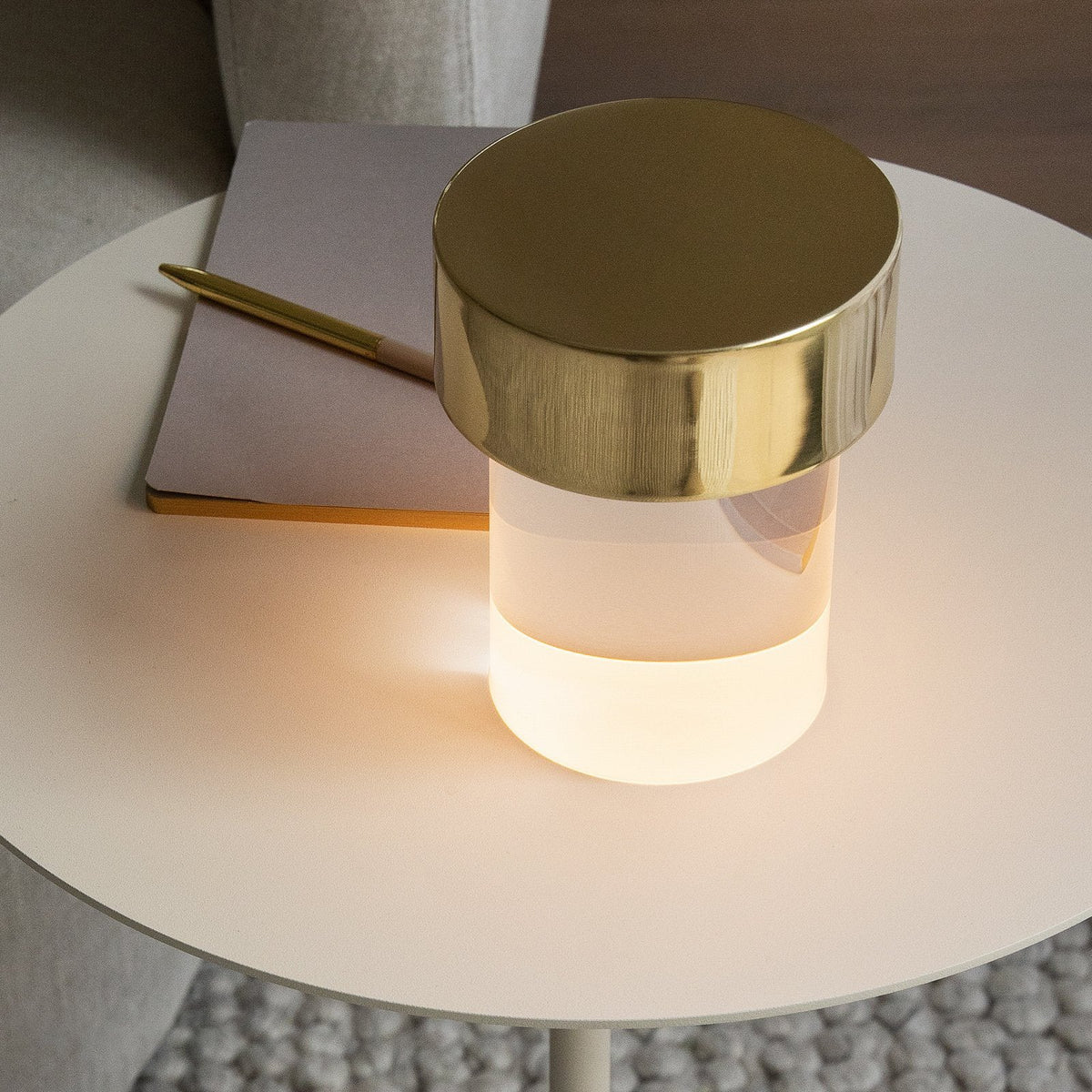 Close view from above of a Clérachic lamp glowing on a round cream side table, beside a notebook and a gold pen.