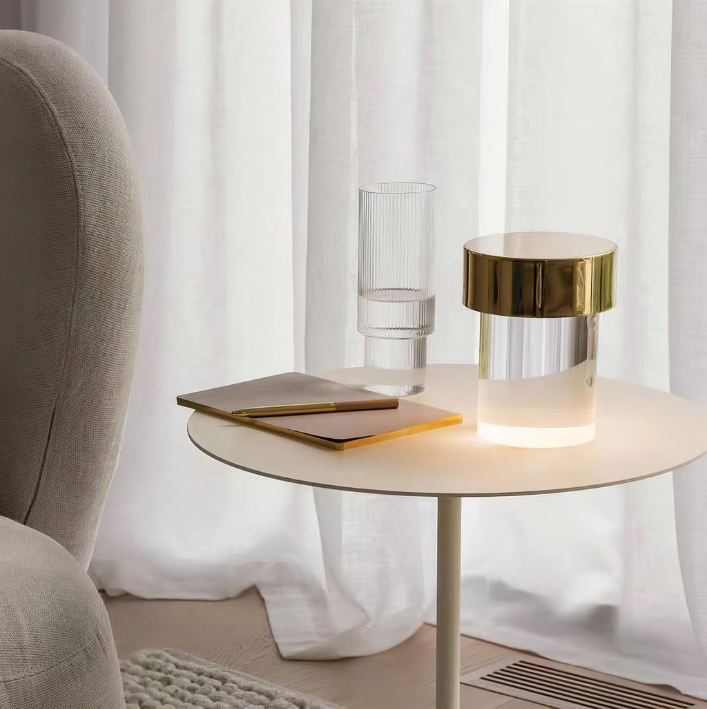 A Clérachic lamp with a gold cap lit on a slim cream side table, with a notebook and a ribbed glass beside sheer white curtains.