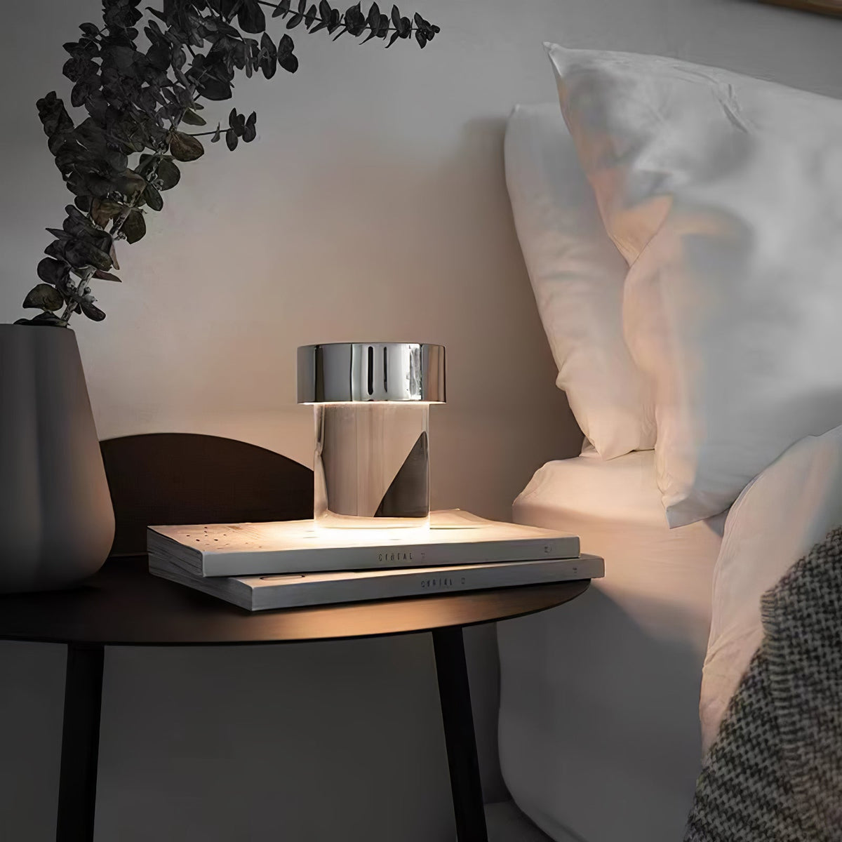 A Clérachic lamp with a chrome cap lit on two stacked books on a dark bedside table, with white pillows and a eucalyptus stem behind.