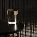 The gold-capped Clérachic lamp on a dark round table in a moody room, its clear glass base catching a thin line of light.