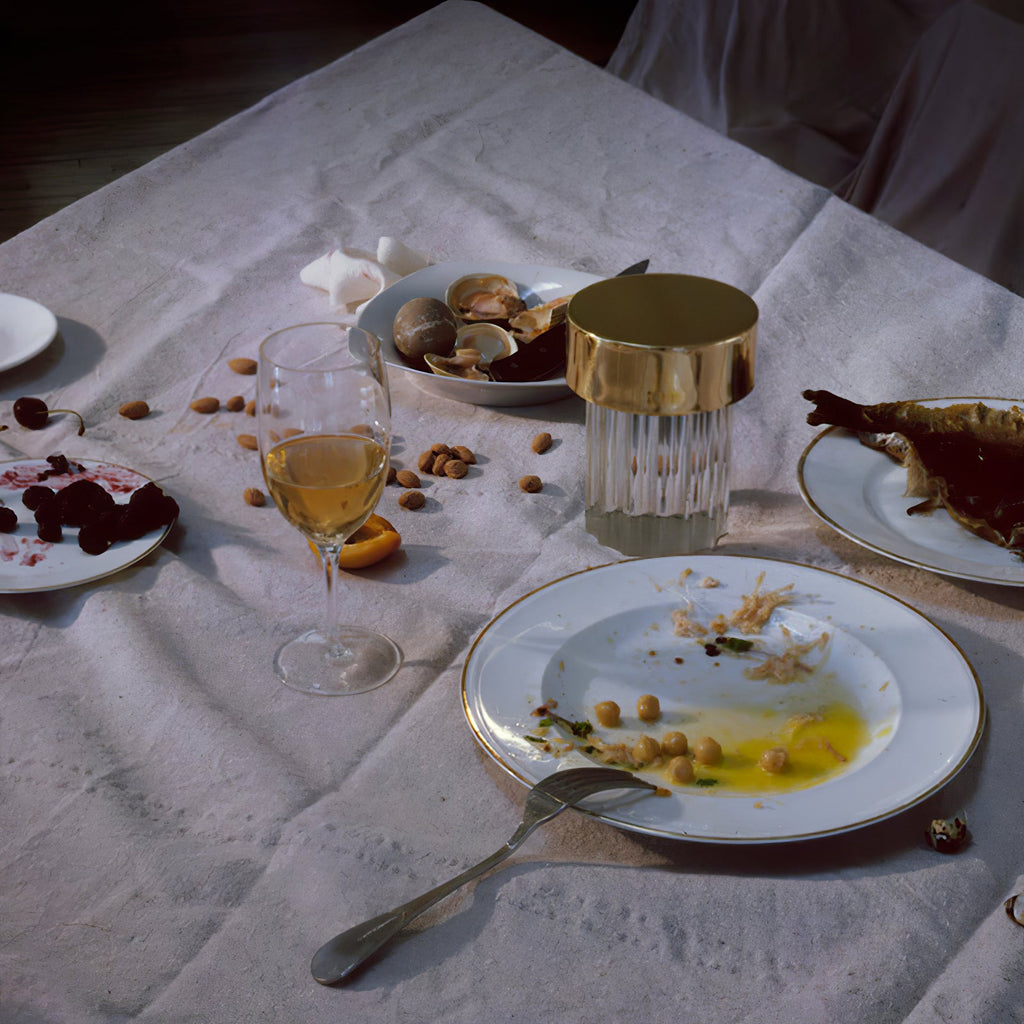 A fluted-glass Clérachic lamp with a gold cap among plates, shells and wine glasses on a rumpled white tablecloth.