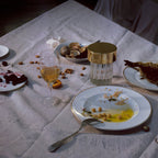 A fluted-glass Clérachic lamp with a gold cap among plates, shells and wine glasses on a rumpled white tablecloth.