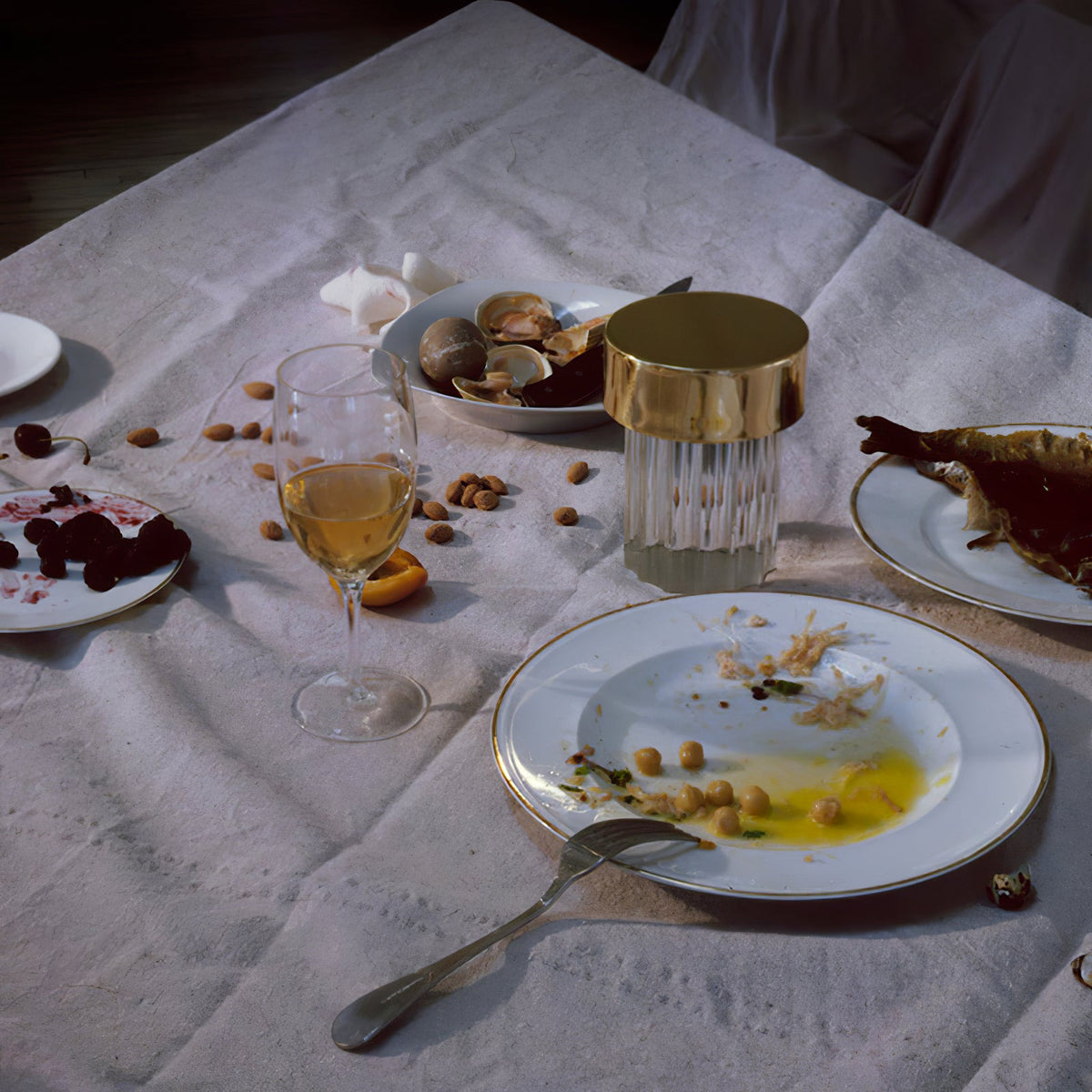A fluted-glass Clérachic lamp with a gold cap among plates, shells and wine glasses on a rumpled white tablecloth.