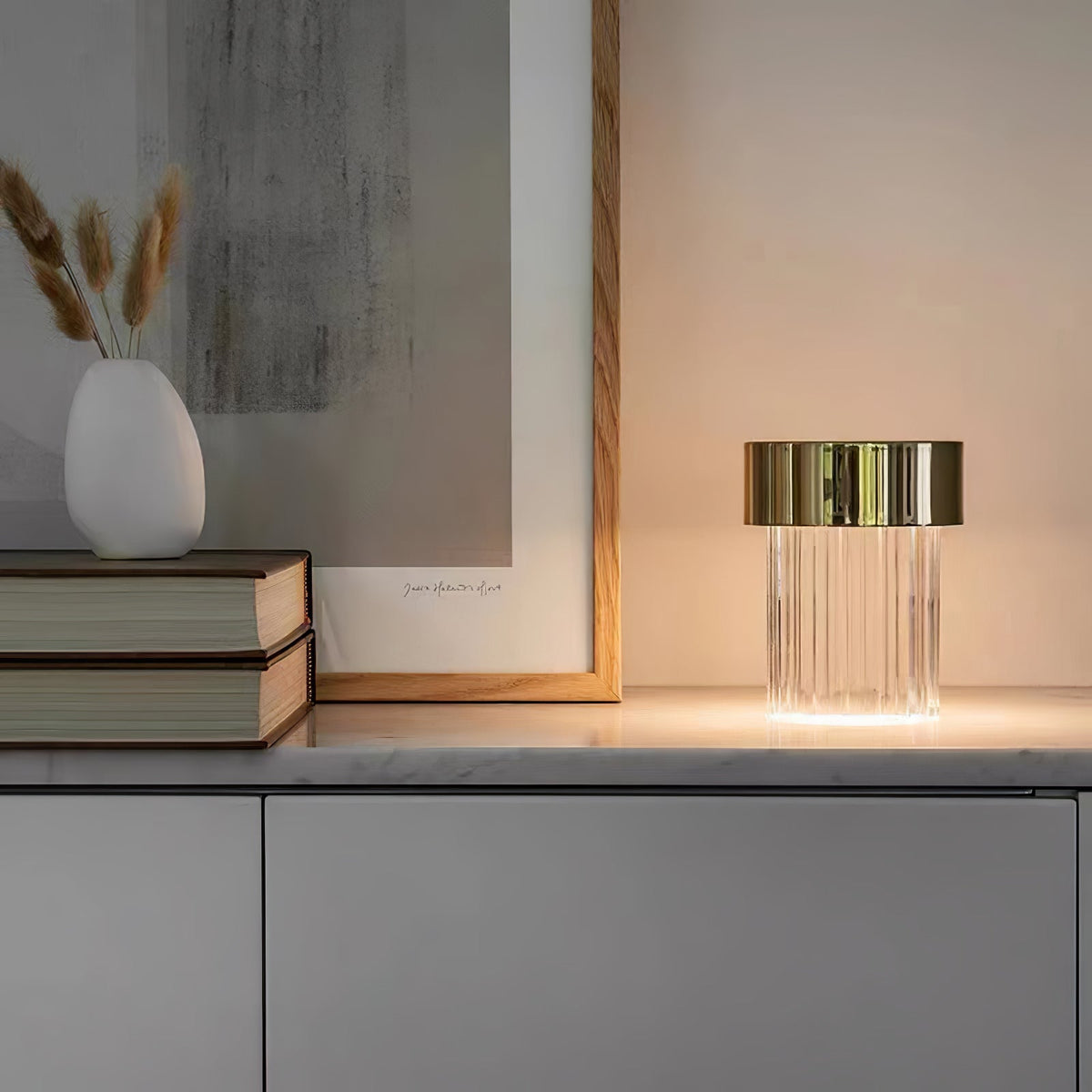A Clérachic lamp with a gold cap and fluted glass glowing on a marble-topped sideboard, beside books, a pale vase and dried grasses.