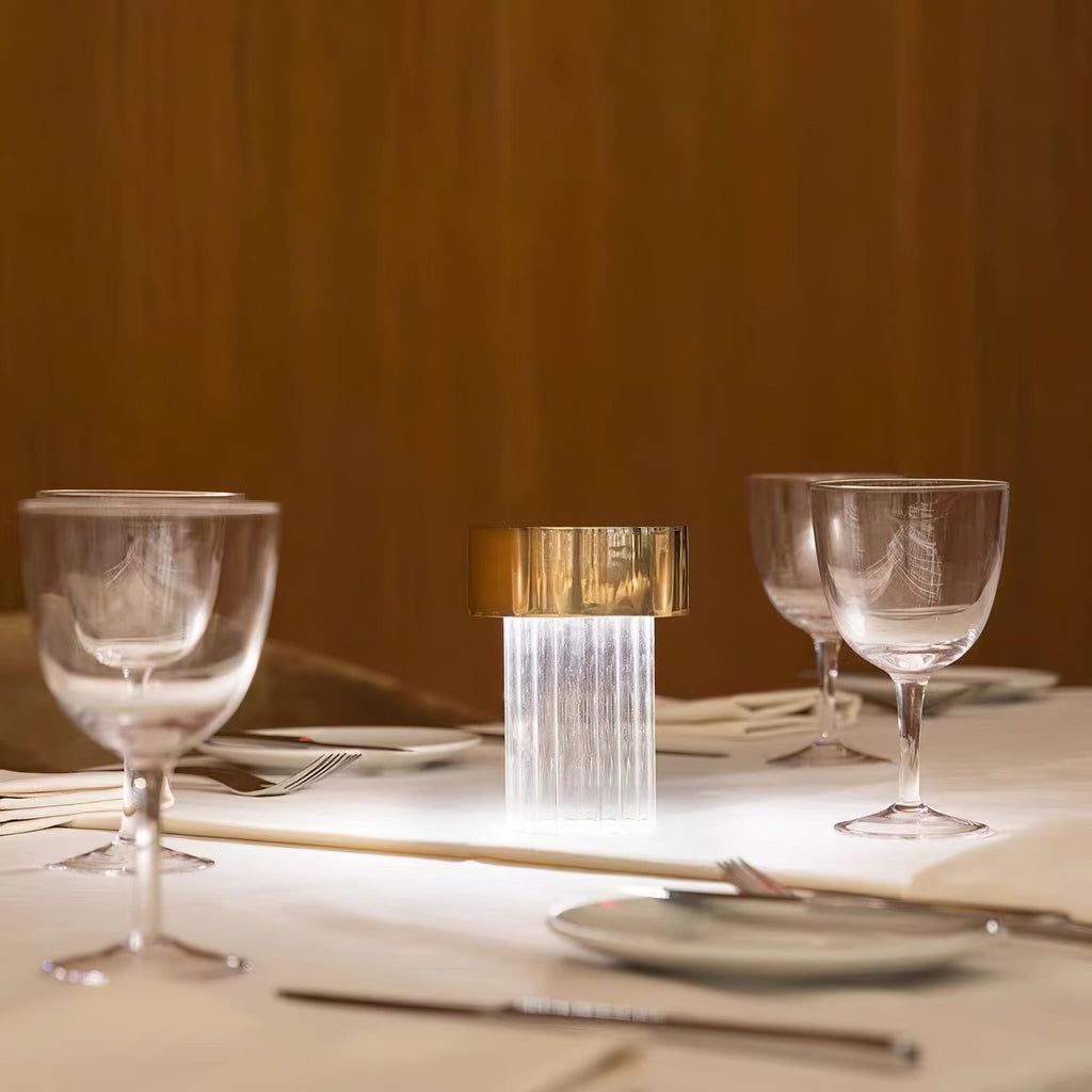 A fluted-glass Clérachic lamp with a gold cap lit on a restaurant table laid with wine glasses, cutlery and a plate.