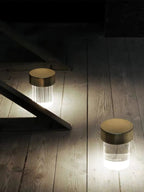 Two Clérachic lamps glowing on a worn wooden floor in a dark room, one fluted and one smooth, both with gold caps.