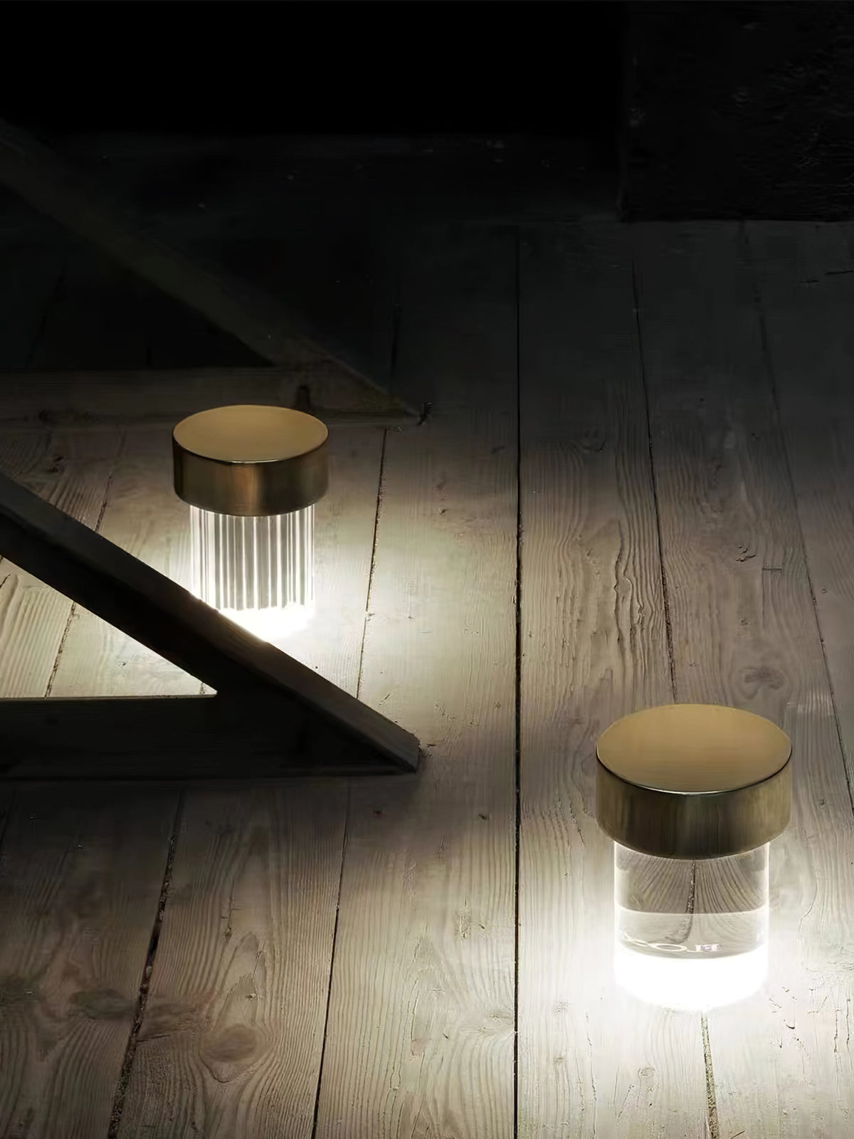 Two Clérachic lamps glowing on a worn wooden floor in a dark room, one fluted and one smooth, both with gold caps.