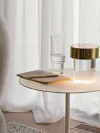 A Clérachic lamp with a gold cap glowing on a cream side table, with a notebook and a ribbed drinking glass beside white curtains.