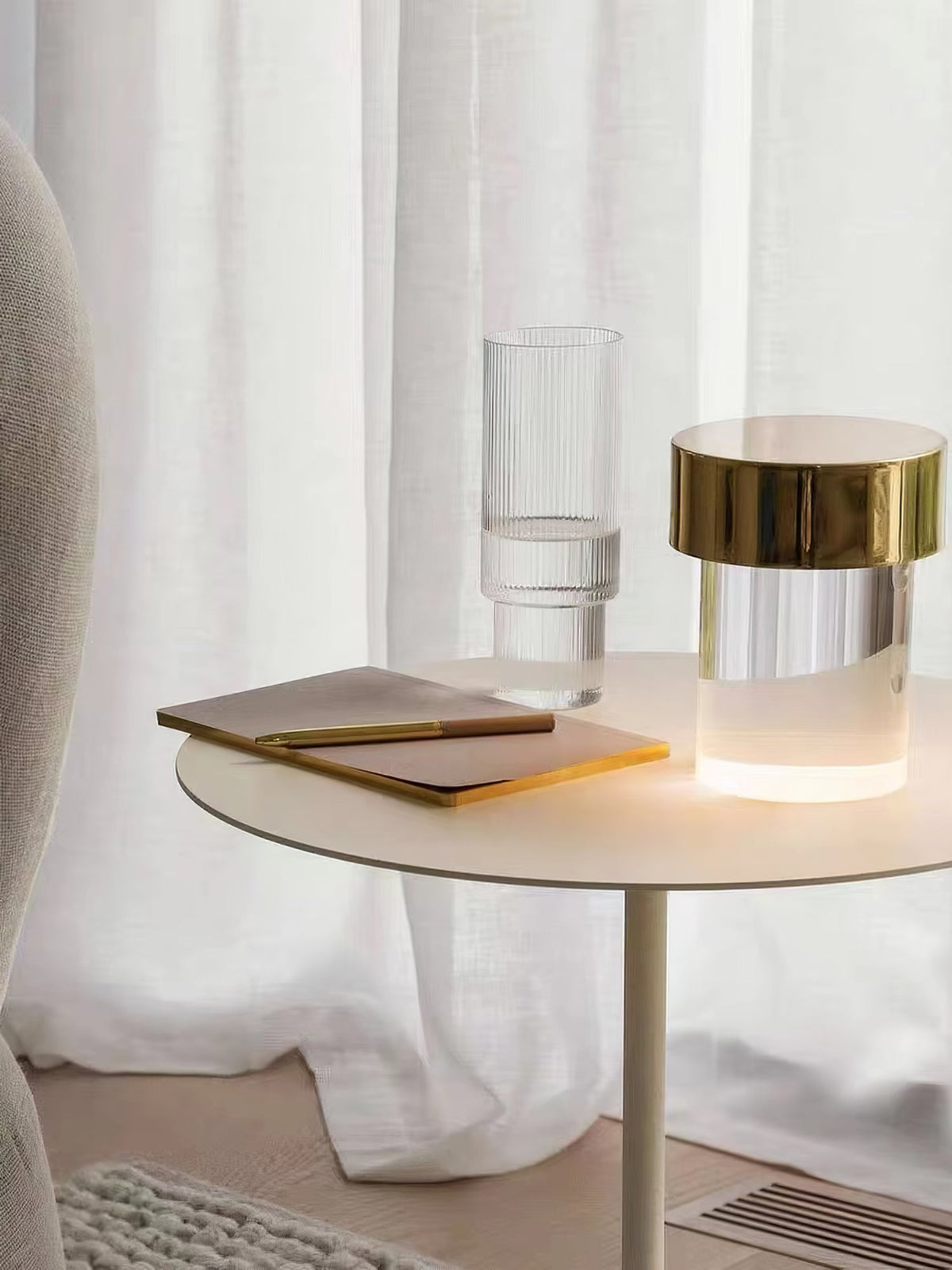 A Clérachic lamp with a gold cap glowing on a cream side table, with a notebook and a ribbed drinking glass beside white curtains.
