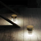 Wide view of two lit Clérachic lamps with gold caps on a pale wooden floor, throwing cool white light across the boards.