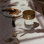 A large gold-capped Clérachic lamp on a white cloth beside a plate of fish, a knife and a few shells, with long shadows.